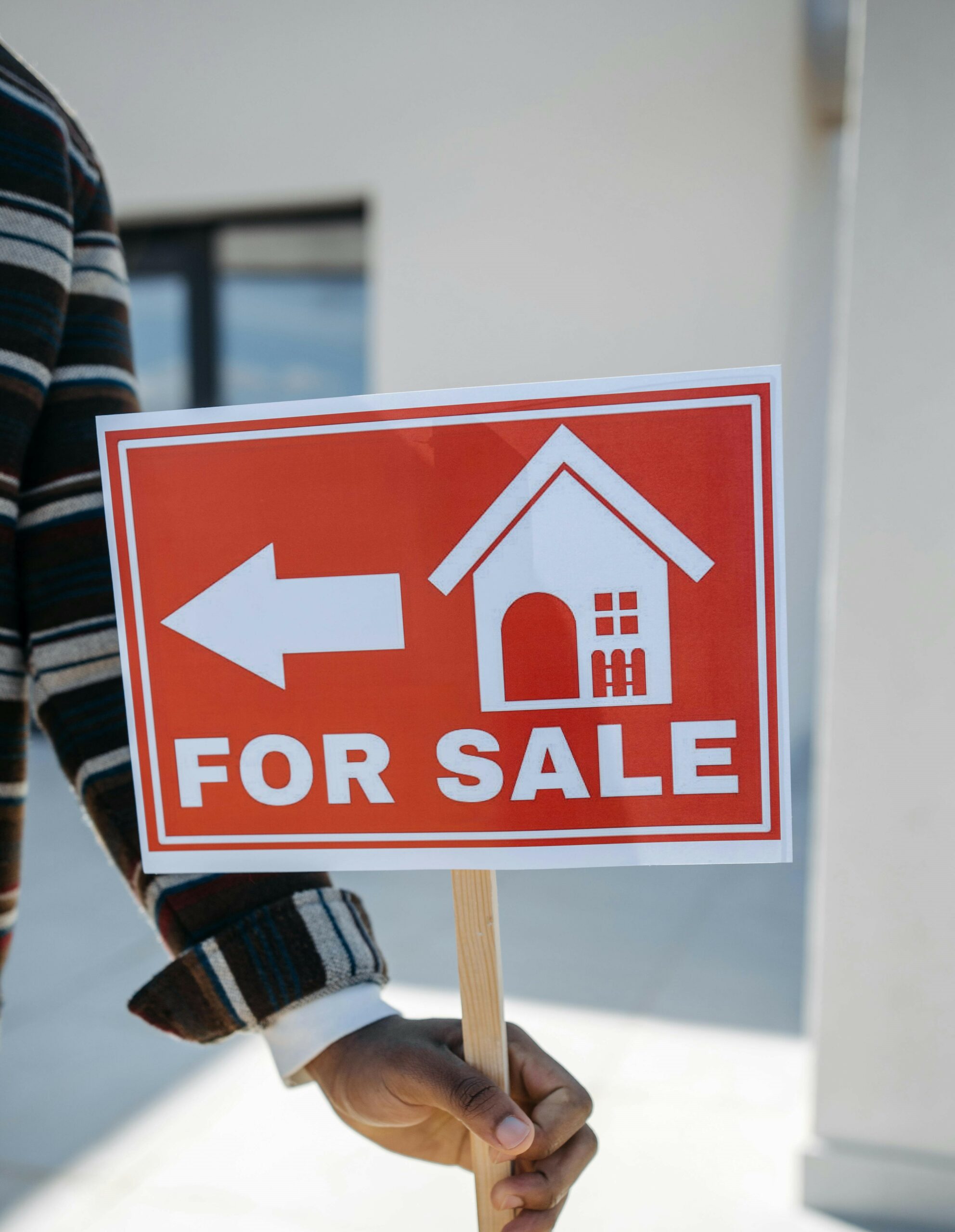 A person in a striped sweater holds a red sign with a white icon of a house and the words "FOR SALE" in uppercase letters, along with a left-facing arrow. The background shows an out-of-focus building exterior.